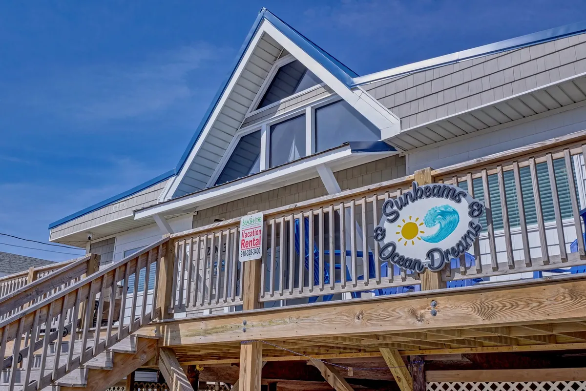This exterior photo centers on a wooden deck adorned with comfortable seating and a 'Sunbeams & Ocean Dreams' sign. The deck enjoys cheerful sunlight and faces the ocean, making it ideal for morning coffee or evening relaxation, as suggested in the section. The architectural details reinforce the connection between outdoor spaces and the inviting entryway.