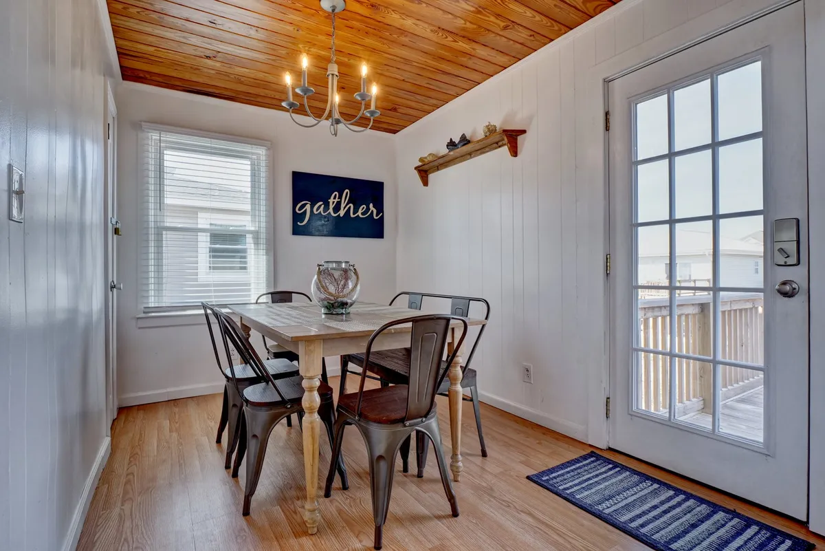A warm, welcoming dining area features a generously sized table beneath a lovely chandelier. A sign reading 'gather' sets a hospitable tone, and the door to the deck suggests easy transitions between indoor and outdoor activities.