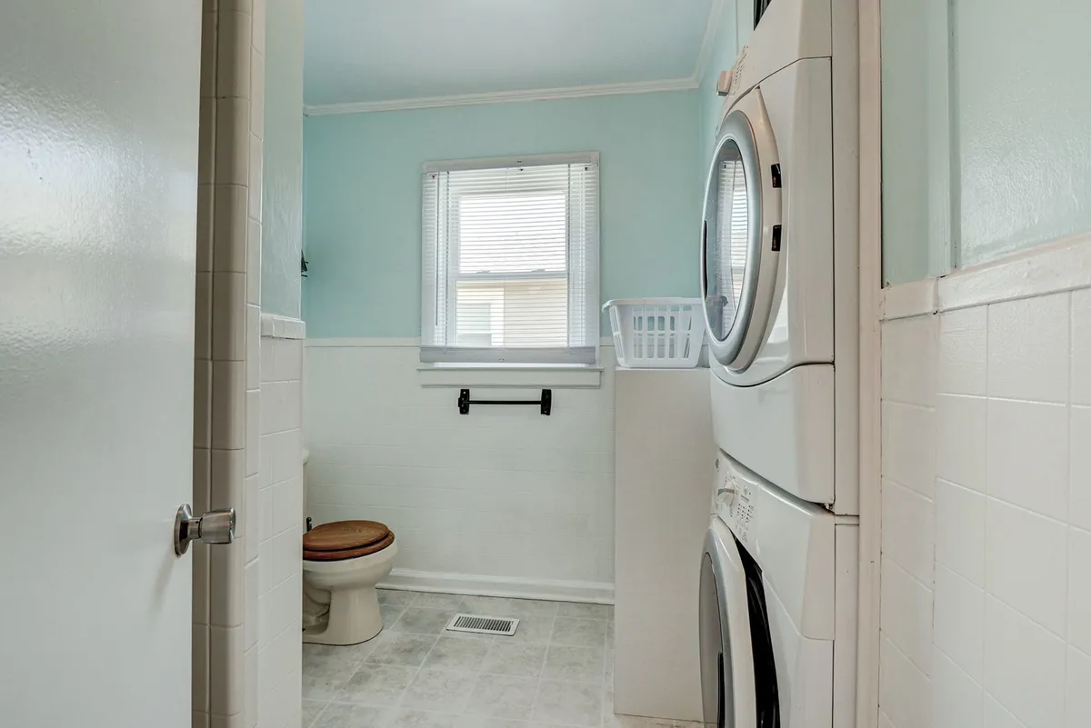 A practical space featuring a stacked washer and dryer, a window with blinds letting in natural light, and a tidy setting with space for laundry essentials.