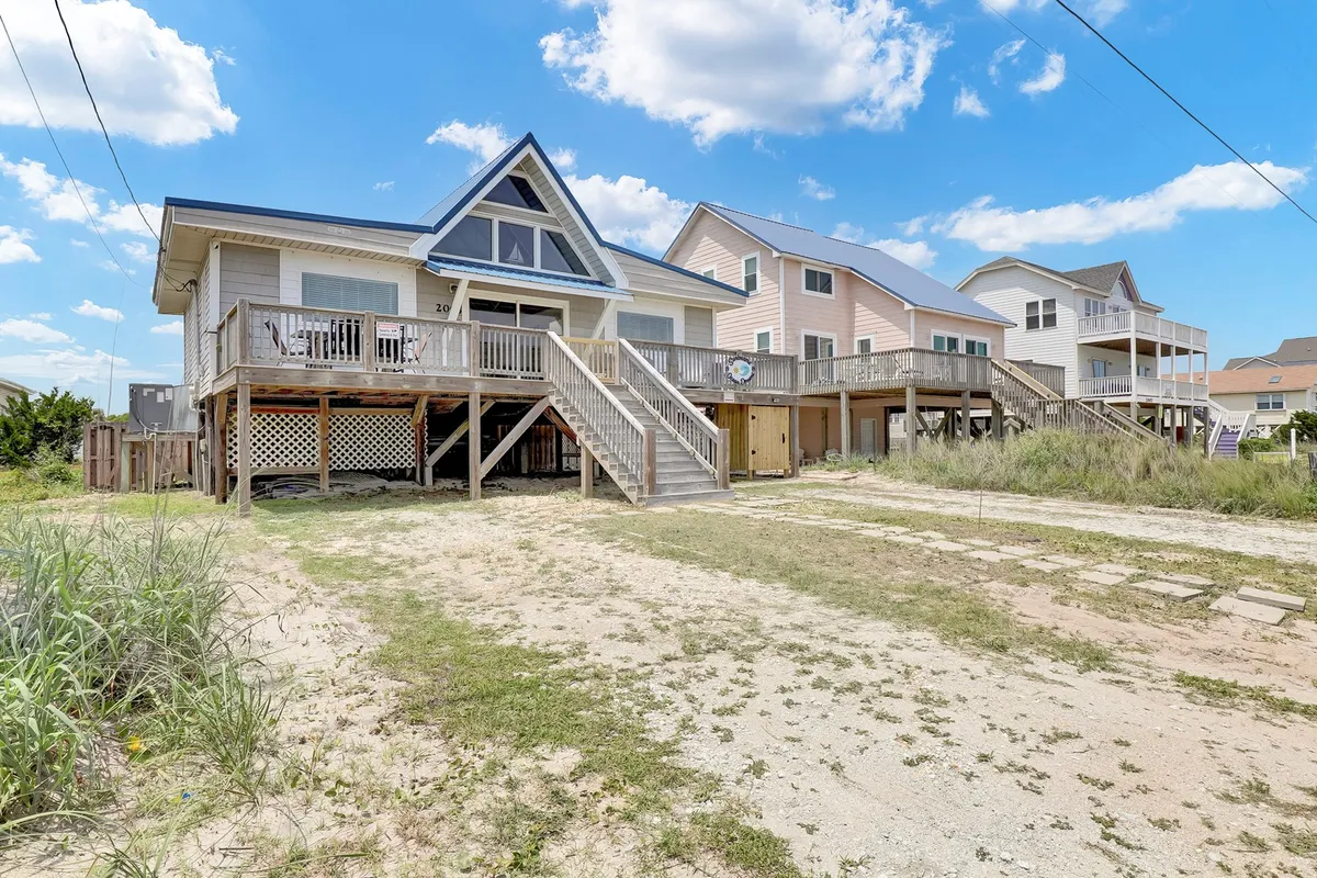 A cheerful exterior view of the beach house featuring a triangular roof, a spacious deck with stairs leading down to a grassy and sandy landscape, and neighboring homes in the background. The deck is clearly visible, positioned to capture sunset views and offer an inviting space for gatherings or relaxation.
