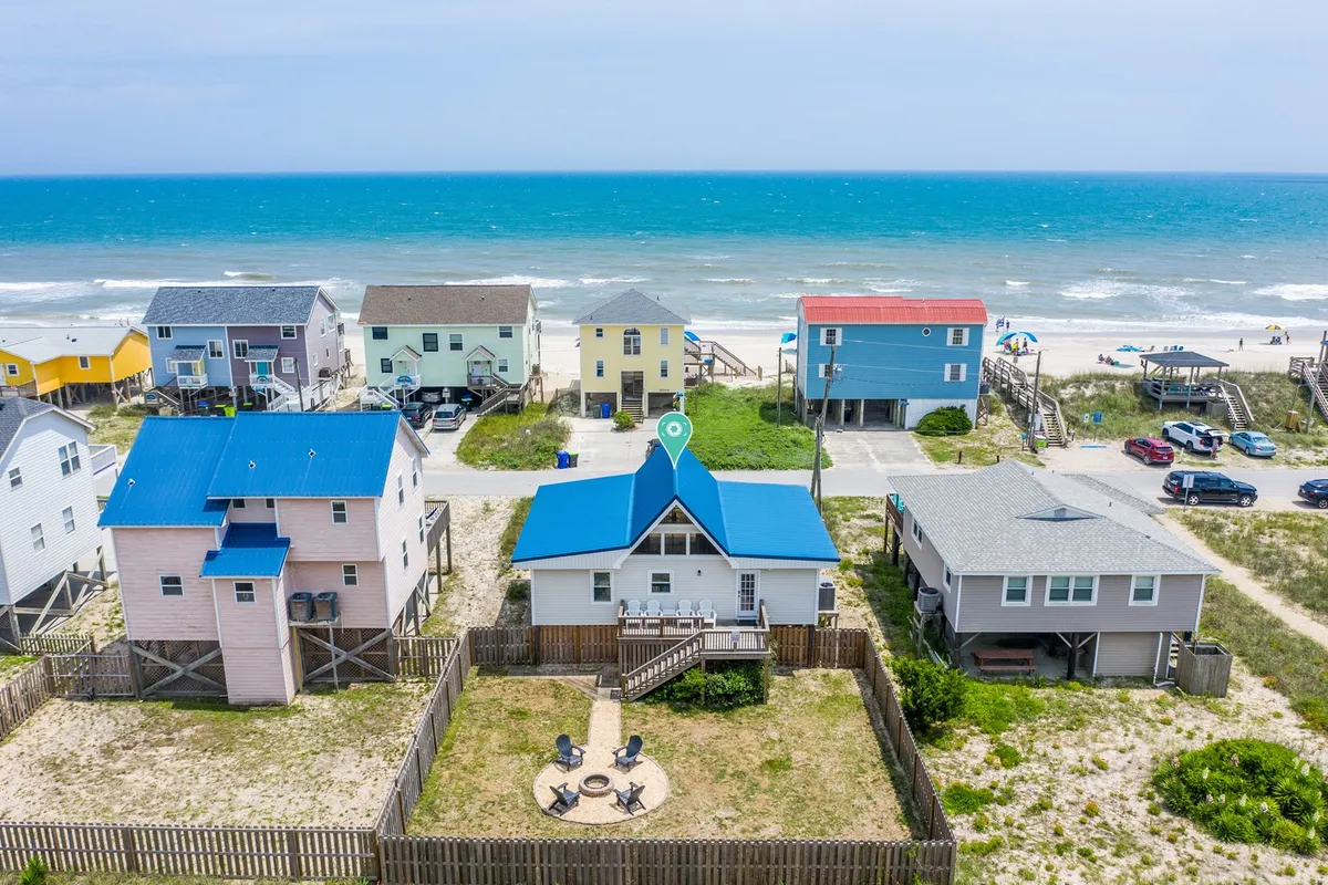 A cheerful beachfront property with a blue roof, open fire pit area surrounded by Adirondack chairs, and colorful neighboring houses. The ocean is visible in the background, and the overall scene conveys a welcoming, social atmosphere ideal for gatherings and relaxation.