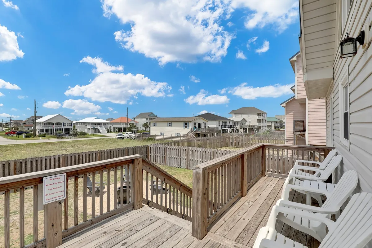 A spacious front deck equipped with white chairs overlooks the neighborhood, with houses in the distance and a blue sky dotted with clouds. The setup is casual and inviting, ideal for relaxing outdoors while enjoying the views and fresh air.