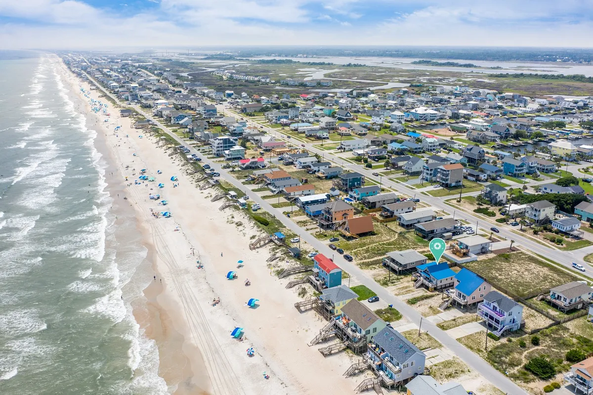 A scenic stretch of sandy beach dotted with umbrellas and beachgoers, bordered by beachfront homes and gentle waves. The wide perspective showcases both the natural beauty and the lively, communal feel of the area.