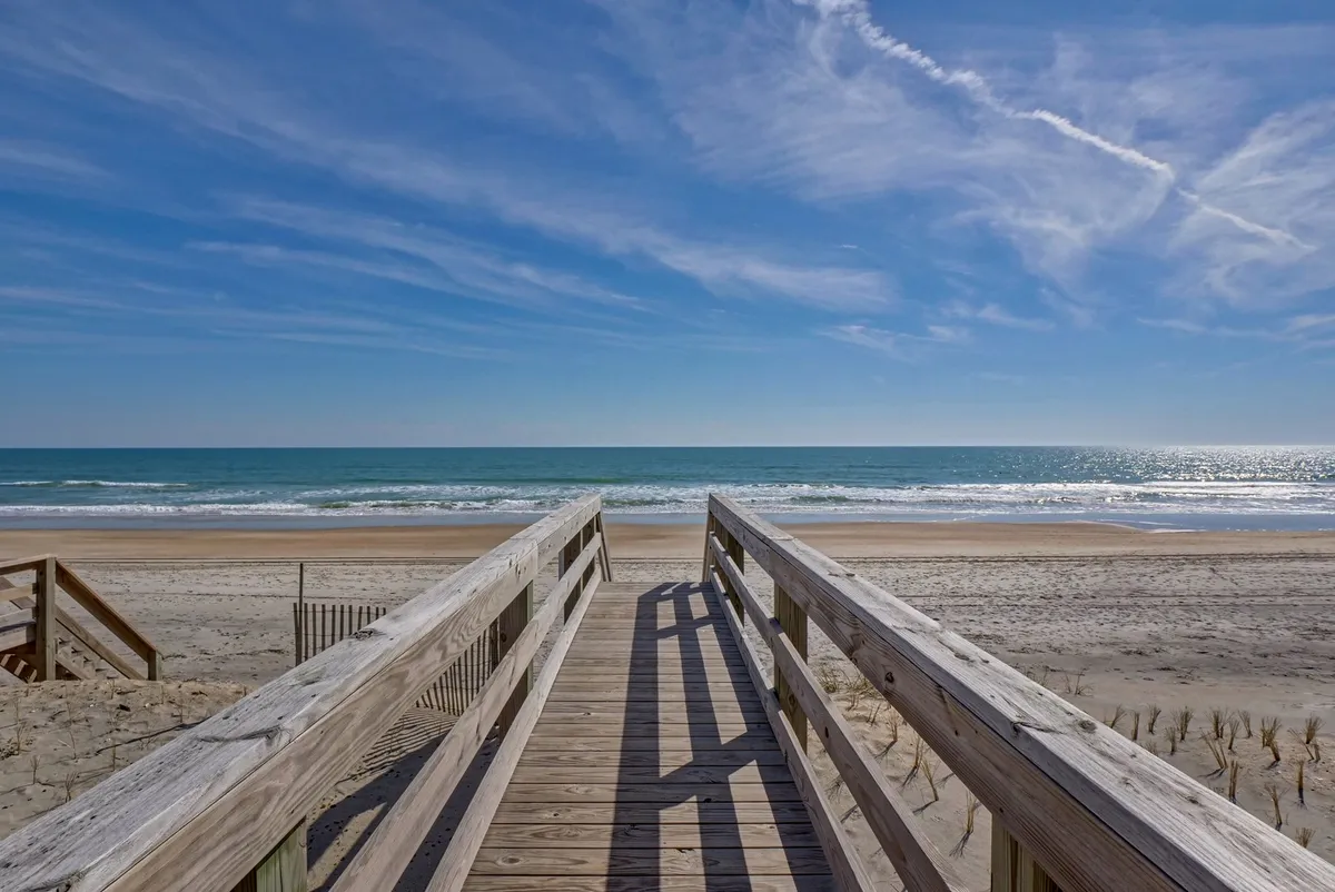 A tranquil wooden walkway leads directly towards the beach, framed by clear skies and views of the ocean horizon. The scene evokes the peaceful start of a day and the inviting possibility of beachfront strolls just beyond the front door.