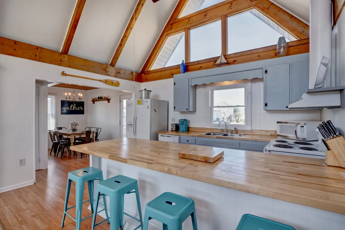 This image showcases an expansive open-concept kitchen with a prominent kitchen island, warm wooden beams drawing the eye upward to vaulted ceilings, large windows filling the space with natural light, and stylish blue cabinetry. Bar stools by the island suggest ample seating for casual meals or conversation.