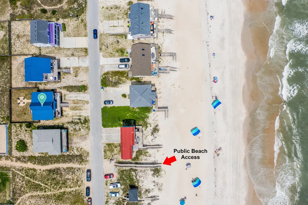 This image captures the public beach access point with colorful beach umbrellas dotting the sandy shoreline and houses lining the coast. The overall scene is lively and inviting, with families and children visible enjoying the sand and surf.