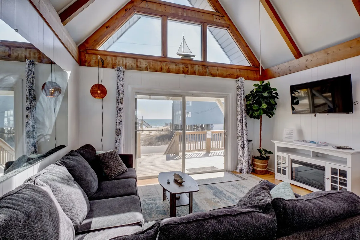 A large sectional sofa takes center stage in the living area, with sliding glass doors letting in light and offering a glimpse of the outdoors. Decorative elements like a sailboat and lush potted plant add to the relaxed, coastal vibe, while the arrangement is ideal for lounging or watching TV.