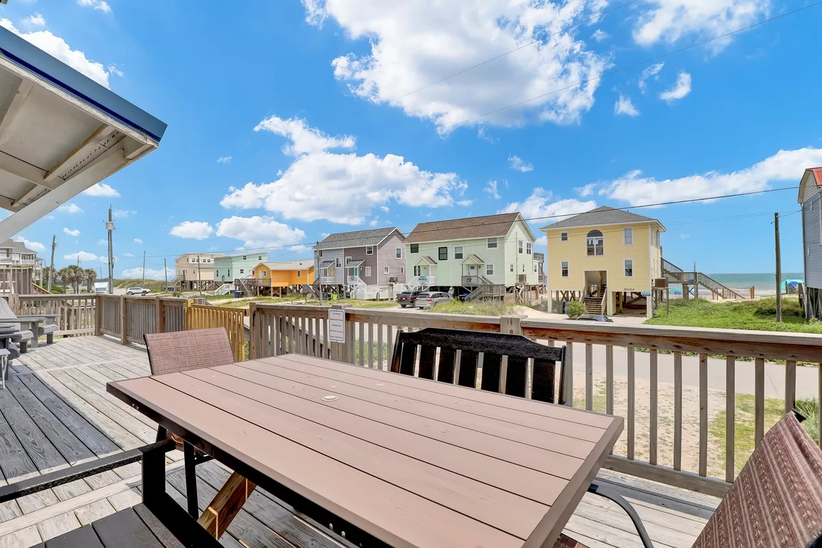 A casual, inviting exterior scene with a deck set with a table and chairs, colorful beach houses nearby, and an open view towards the ocean. The setting clearly conveys a relaxed space for outdoor dining, socializing, and enjoying sunsets over the water.