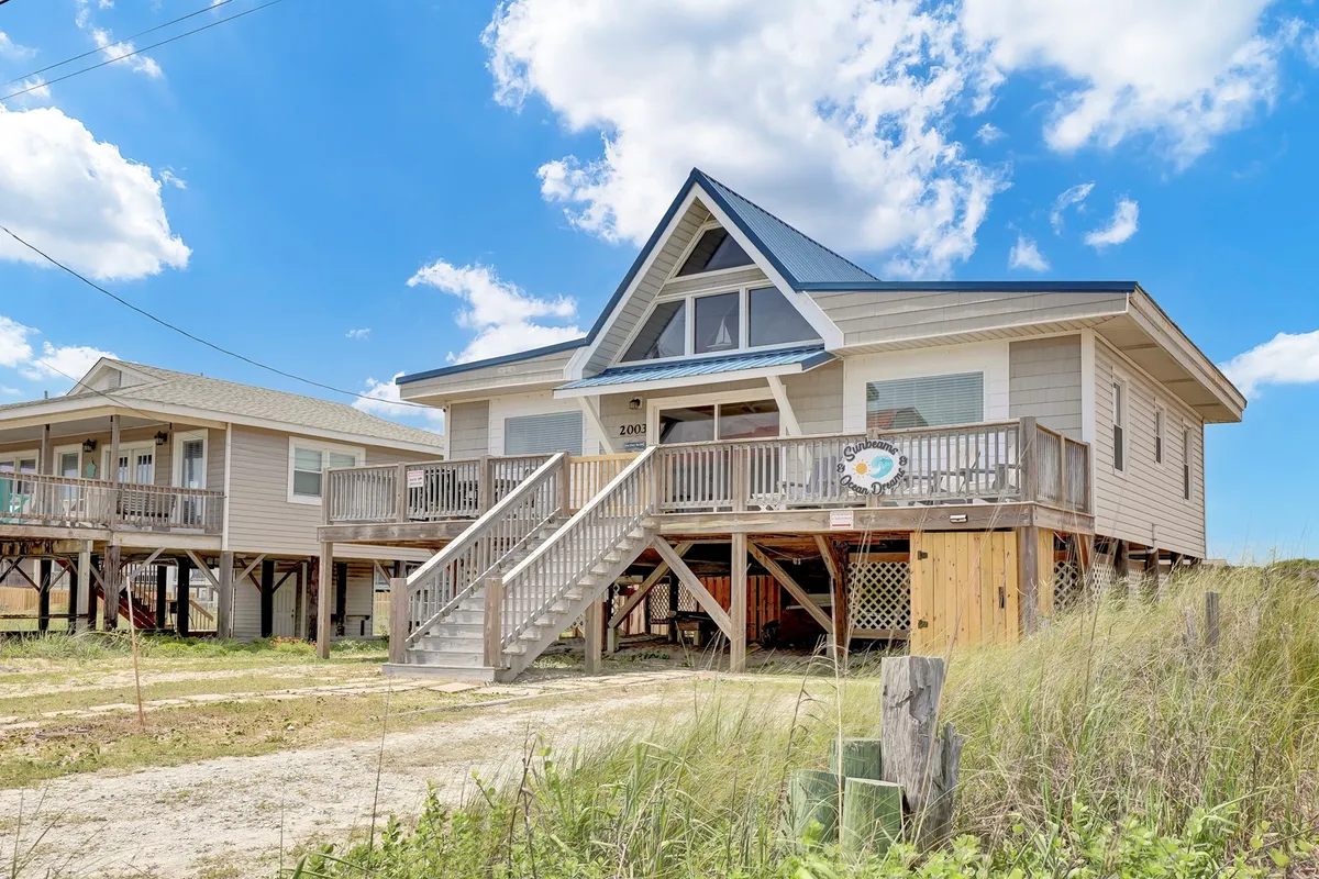 This exterior image showcases the large front porch of the beach house, complete with a welcoming sign, A-frame roof, and inviting atmosphere. Adirondack chairs are visible, perfectly capturing the emphasis on relaxing outdoor living. The orientation of the porch suggests a view toward the ocean, and the bright daylight highlights the oversized space and tranquil mood.