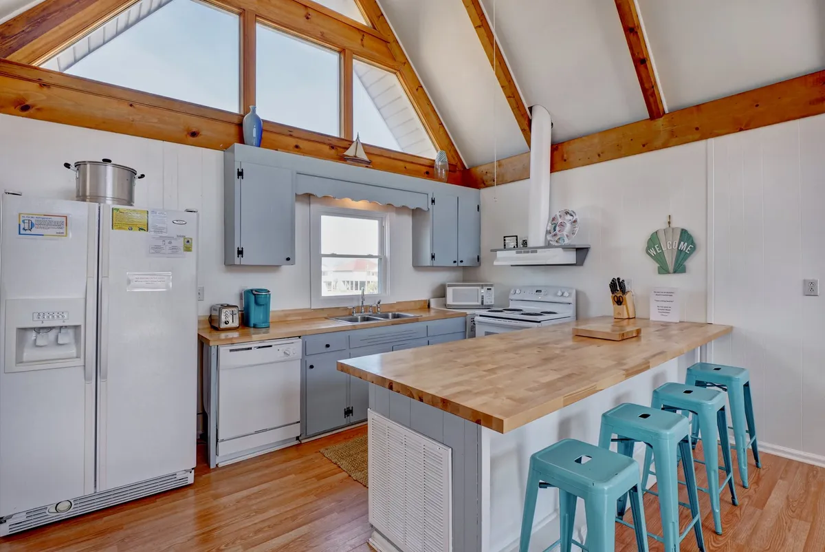 A warm, sunlit kitchen featuring wooden countertops, blue cabinetry, large windows, and bar stools. The space is designed for cooking, entertaining, and enjoying shared meals, providing a practical yet welcoming environment.