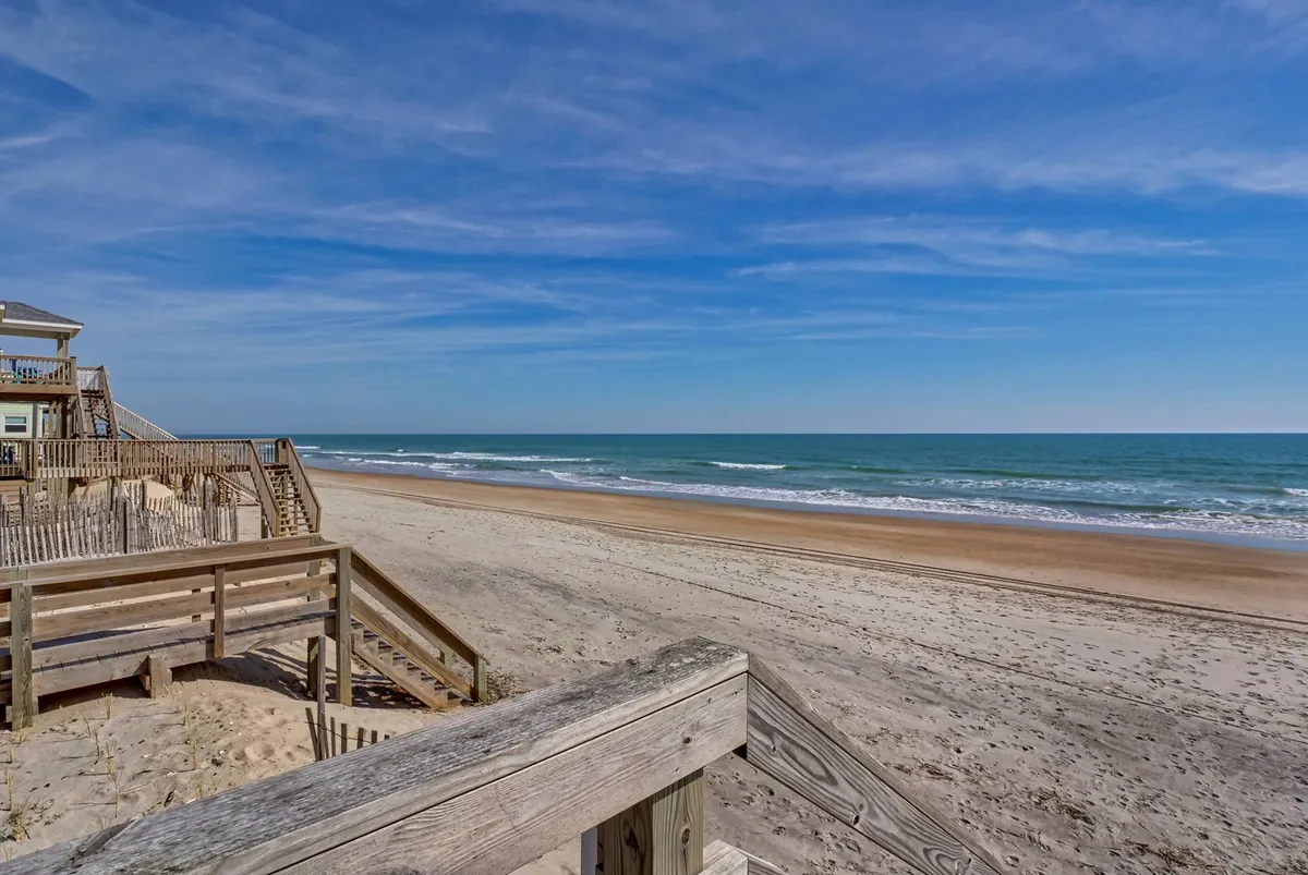 A bright, inviting beach scene featuring a stretch of sandy shore, gentle ocean waves, and a charming wooden boardwalk leading to the water. People can be seen enjoying beach activities such as sunbathing and walking along the shore, capturing the relaxed vacation atmosphere.
