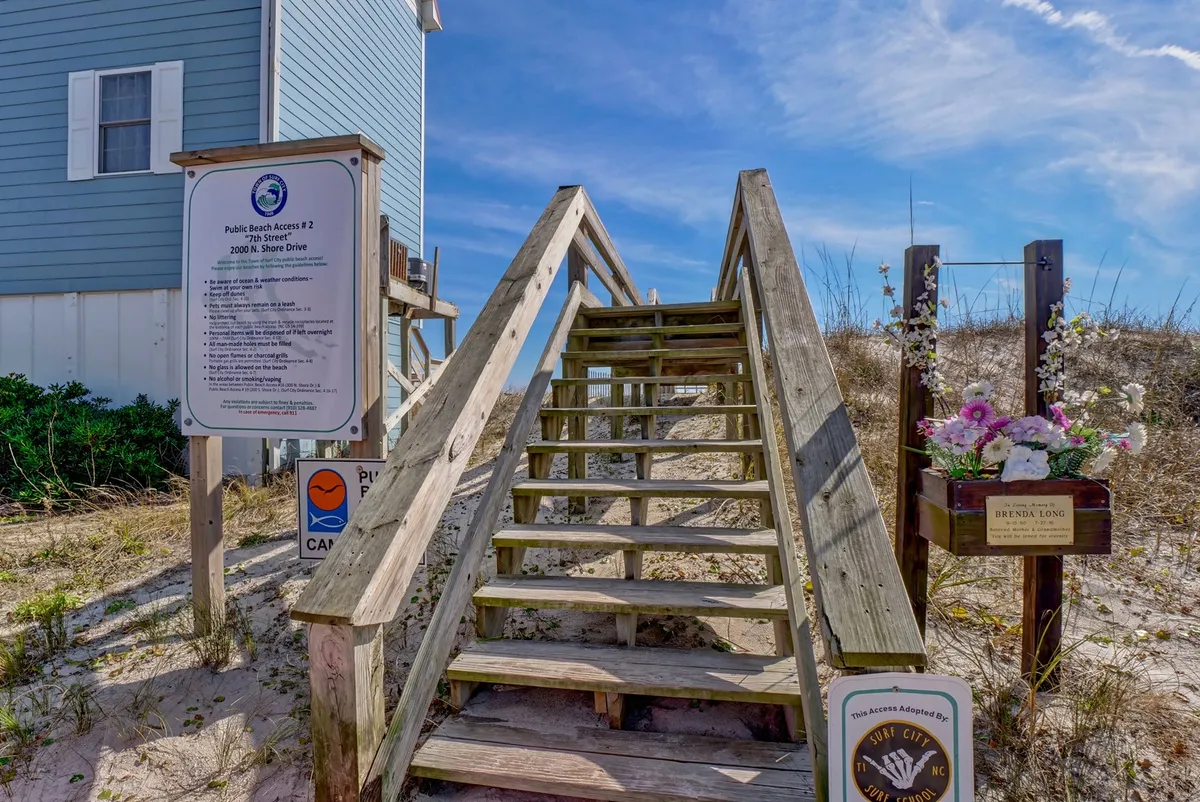 A wooden staircase leads directly to the beach, flanked by a beach access sign and a decorative flower box. The steps descend toward the sand, evoking the anticipation of a beach day.